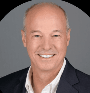 Smiling bald man in a white shirt and dark blazer against a grey background.